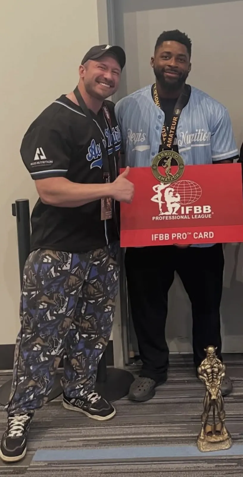 David DeMesquita with his athlete holding an IFBB Pro card and trophy